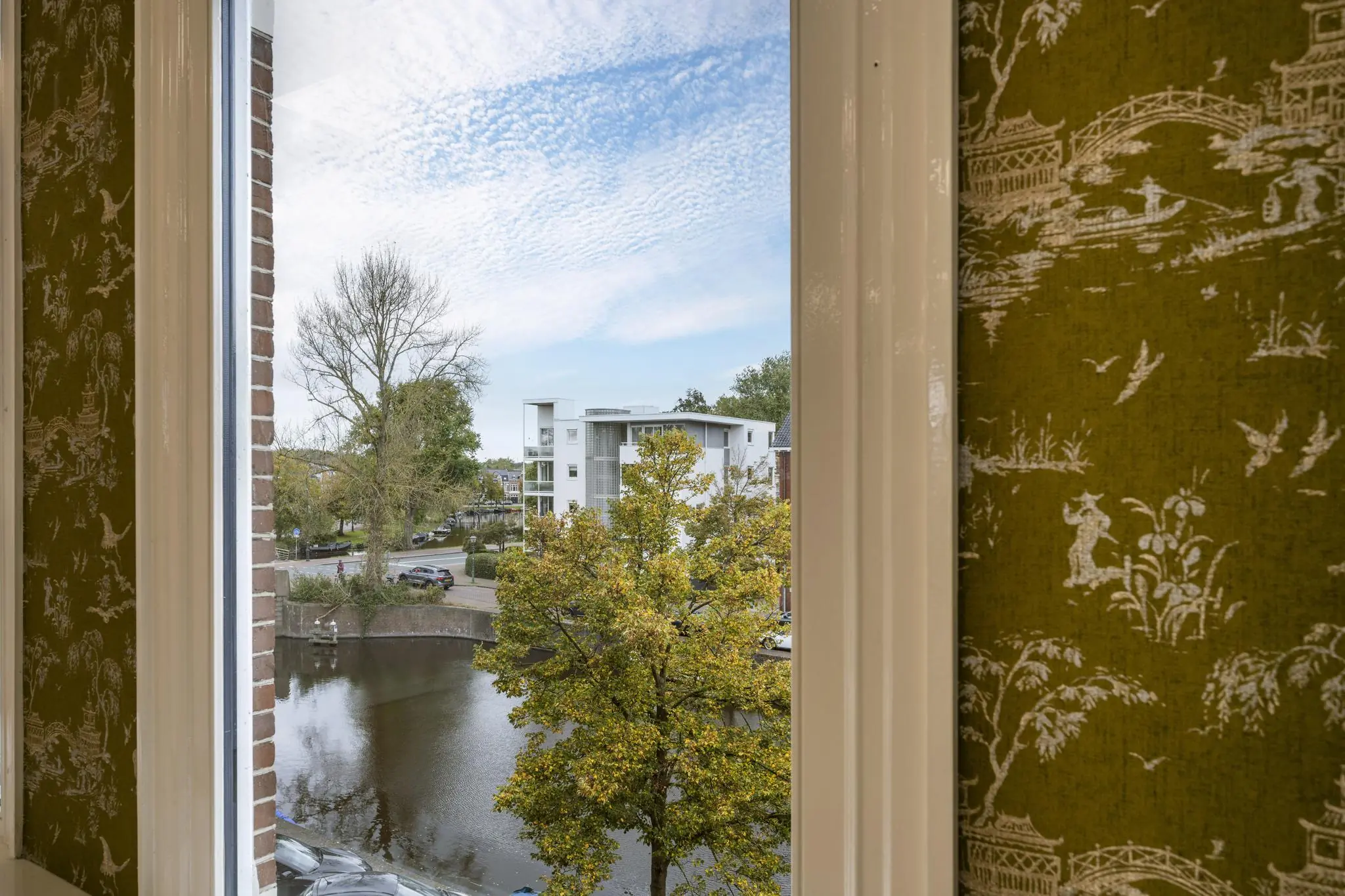 Uitzicht vanuit een raam op de Nieuwe Gracht met bomen, water en moderne witte gebouwen.